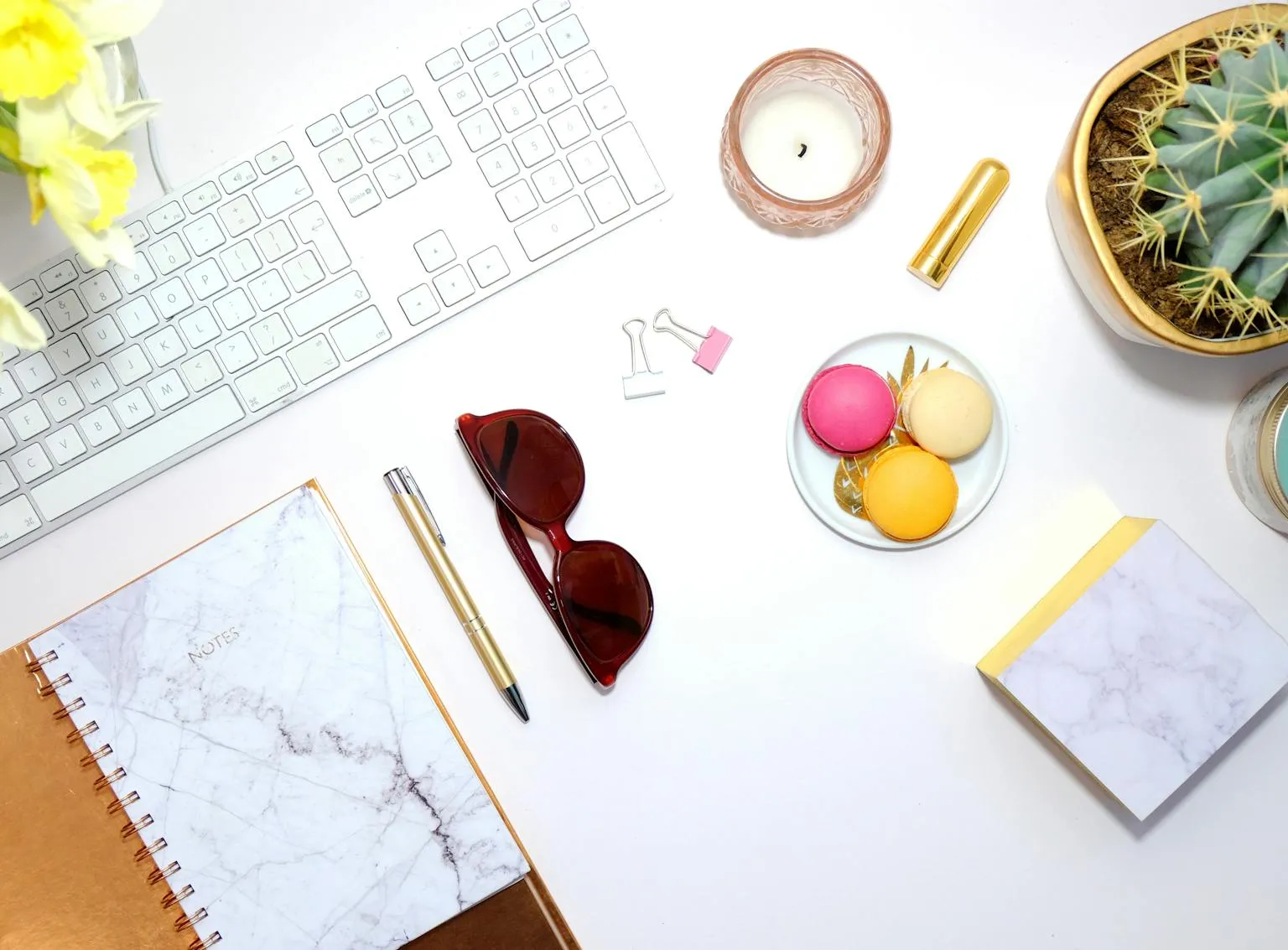 A writing desk from above with a keyboard, notebook, pen, sunglasses, a candle and macarons